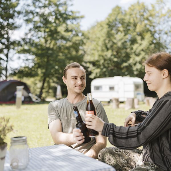 Zwei Personen sitzen entspannt am Campingplatz und stoßen mit einem Getränk an